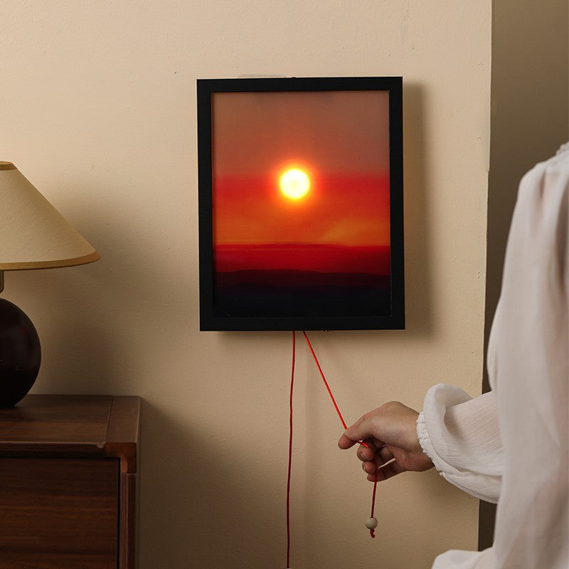 Framed artwork of a sunset with a hand pulling on a string, next to a lamp on a wooden table.